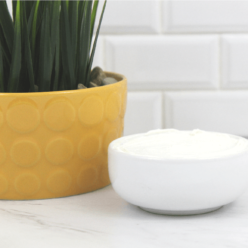 Body creme in a white bowl sitting next to a yellow plant pot filled with a grass plant